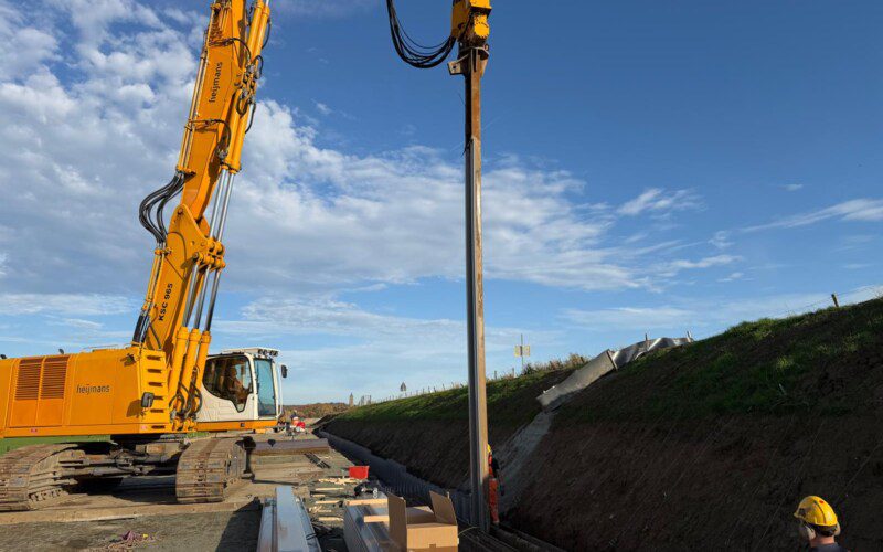 Kraan brengt een Prolock Epsilon kunststof damwand in met behulp van een stalen moederplank tijdens dijkversterkingswerkzaamheden, begeleid door waterbouwspecialisten op de bouwplaats.